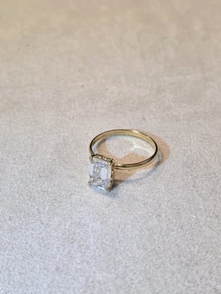 Gold engagement ring with rectangular emerald cut diamond solitaire on cream fabric background